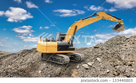 An excavator is moving dirt at a construction site with a clear blue sky and scattered clouds overhead. Piles of gravel and rocks are visible around the machine An excavator is moving dirt at a construction site with a clear blue sky and scattered clouds overhead. Piles of gravel and rocks are visible around the machine 133440803