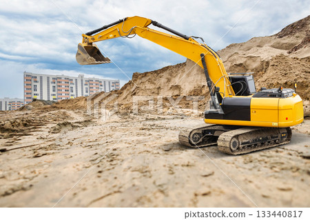 A powerful crawler excavator is seen digging on a sandy construction site, surrounded by tall residential buildings under a cloudy sky 133440817