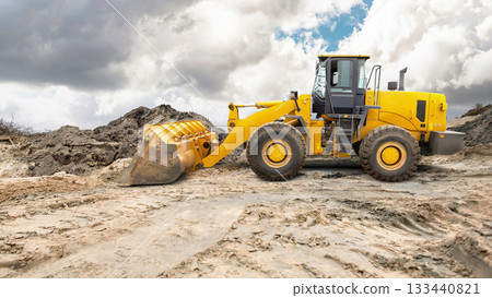 Heavy machinery moves dirt at a construction site. A yellow loader uses its bucket to lift soil. Fluffy clouds fill the sky, creating an overcast afternoon scene Heavy machinery moves dirt at a construction site. A yellow loader uses its bucket to lift soil. Fluffy clouds fill the sky, creating an overcast afternoon scene 133440821