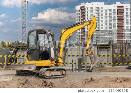 Mini excavator is actively digging at a construction site while a modern skyline is visible under a blue sky with scattered clouds Mini excavator is actively digging at a construction site while a modern skyline is visible under a blue sky with scattered clouds 133440838