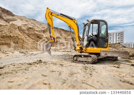An excavator works to clear sand at a construction site surrounded by tall buildings under a cloudy sky, showcasing urban development activities 133440858