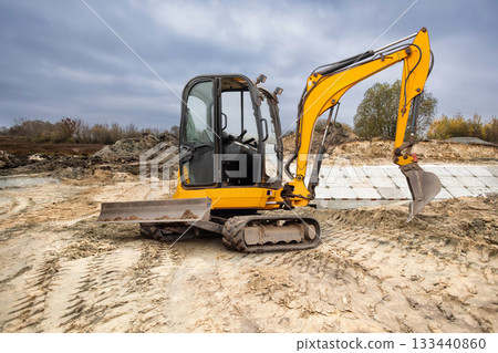 A yellow excavator is positioned on a construction site, preparing to dig into the sandy ground with cloudy skies overhead A yellow excavator is positioned on a construction site, preparing to dig into the sandy ground with cloudy skies overhead 133440860