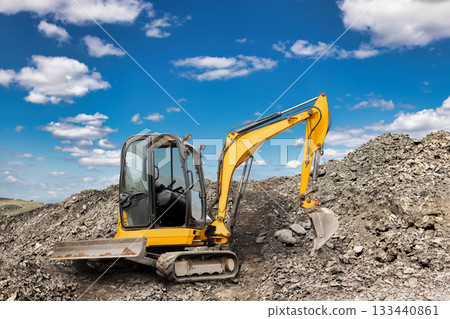 A yellow excavator is digging into a mound of dirt at a construction site. The sun shines brightly, casting shadows on the rugged terrain 133440861