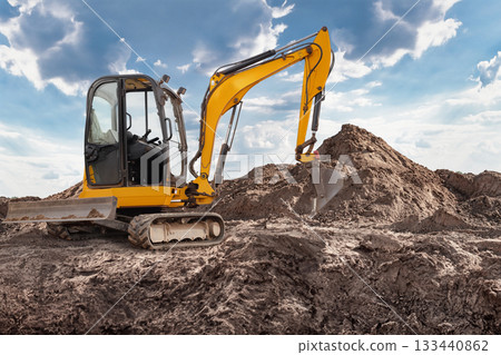 Mini excavator is digging and moving earth at a construction site with a large dirt pile nearby, showcasing industrial activity in progress 133440862