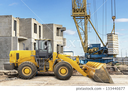 Yellow loader and crane operate on a construction site, moving heavy materials among partially built structures under a clear sky 133440877
