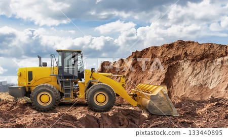 A yellow loader operates on a large dirt mound at a construction site. The machine is moving soil with its bucket against a cloudy backdrop 133440895