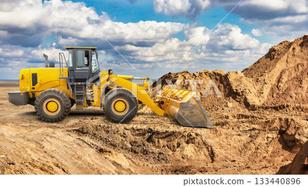 A large yellow wheeled bulldozer or loader moves a pile of dirt at a construction site under a cloudy sky, showcasing the activity of land clearing and preparation A large yellow wheeled bulldozer or loader moves a pile of dirt at a construction site under a cloudy sky, showcasing the activity of land clearing and preparation 133440896