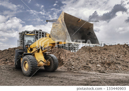 A large bulldozer loader lifts a pile of dirt on a construction site surrounded by buildings and cranes under a cloudy sky during the day A large bulldozer loader lifts a pile of dirt on a construction site surrounded by buildings and cranes under a cloudy sky during the day 133440903