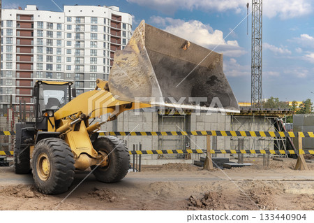 A yellow loader lifts dirt at a busy construction site surrounded by an urban landscape with tall buildings on a clear day 133440904