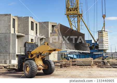 Heavy machinery is actively engaged on a construction site, with a loader moving dirt and a crane positioned to lift materials against a blue sky 133440905