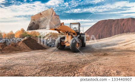 A large wheel loader removes soil from a site surrounded by hills. The sky is clear, and the trees are painted in autumn colors, heralding the arrival of autumn 133440907