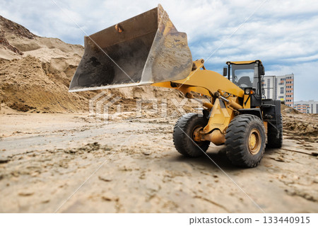 A loader is seen lifting a heap of dirt at a construction site in a city, surrounded by tall buildings and a cloudy sky during the day 133440915