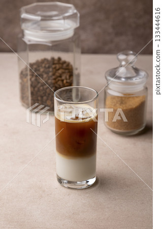 Coffee with coconut milk and ice cubes in a tall glass. Traditional non-alcoholic cold drink. In the background, a jar of coffee and a jar of sugar. Selective focus, close-up. Coffee with coconut milk and ice cubes in a tall glass. Traditional non-alcoholic cold drink. In the background, a jar of coffee and a jar of sugar. Selective focus, close-up. 133444616
