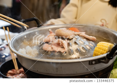 Sliced meat and vegetables cook in a metal grill pot filled with broth and steam, while the center grill browns the meat. 133446375