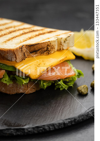 Grilled salmon and cheddar sandwich with lettuce on slate board, lemon and capers 133447351