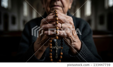 Wrinkled hands of an elderly Catholic woman hold a wooden rosary with a metal crucifix. The scene captures a moment of prayer and contemplation in a serene church environment. 133447739