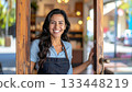 Happy woman wearing apron opening a door, inviting people into her shop. 133448219
