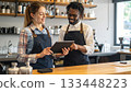 Two smiling baristas collaborating and using digital tablet at the coffee shop counter. 133448223