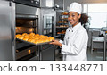 Young woman chef smiling, holding a tray of baked goods in professional kitchen. 133448771