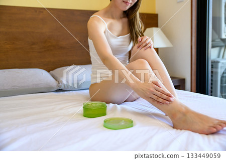 A young, slender woman in underwear applying body gel to her legs in bed. Foot care. Daytime color. 133449059