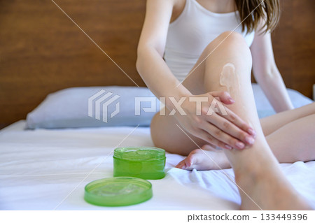 A young slender woman in underwear applying body gel to her legs in bed, foot care, front view, morning color 133449396