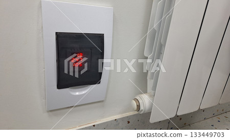 Digital thermostat display with temperature settings on wall panel near radiator 133449703