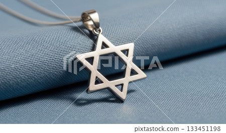 A macro shot of a silver Star of David necklace on a blue textured background. This image represents faith, Judaism, and cultural identity. Perfect for religious or spiritual themes. 133451198