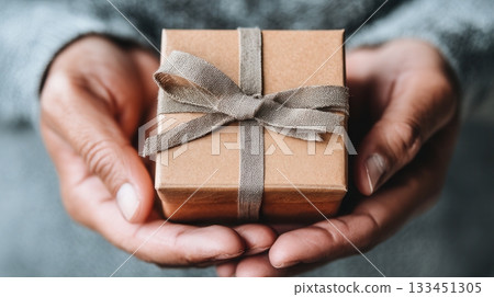 Close-up of hands holding a small kraft gift box with a ribbon. This image evokes warmth, love, and the joy of giving. Ideal for holidays, birthdays, or special occasions. 133451305
