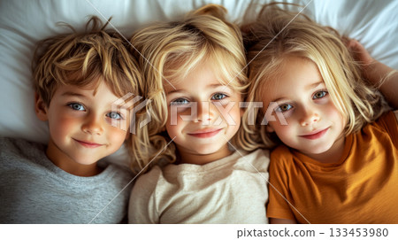 Three children with blond hair lie side by side, smiling on a bed. Concept of happiness, innocence, and togetherness. 133453980