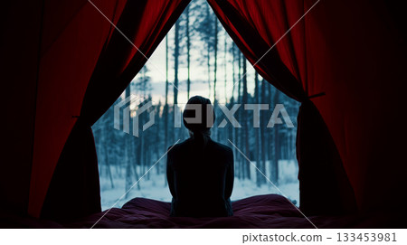 A silhouette of a person is sitting inside a tent, looking out at a snowy forest through red curtains. Concept of solitude, contemplation, and nature. 133453981
