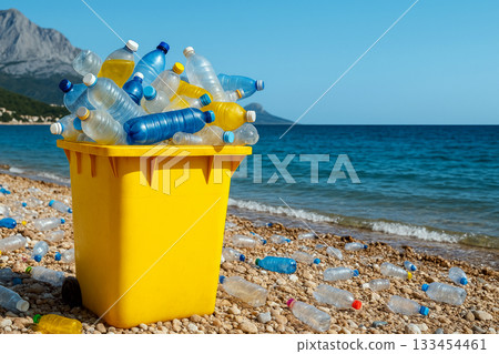 Yellow trash bin filled with plastic bottles on a pebble beach, with blue sea in the background. Concept of pollution and environmental awareness. Yellow trash bin filled with plastic bottles on a pebble beach, with blue sea in the background. Concept of pollution and environmental awareness. 133454461