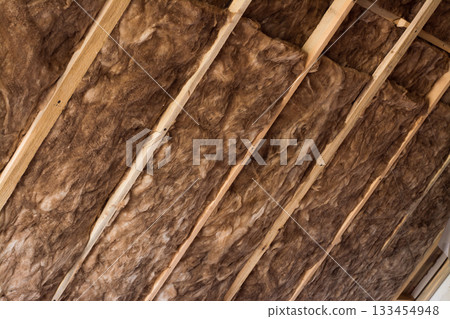 Brown glass wool in a wooden frame on a inclined wall near the wooden ceiling in a private house. Warming the house with fiberglass. 133454948