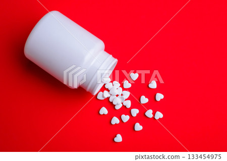 Small white pills in the shape of hearts fell from white medicine bottle laying on red colorful background. Treatment of heart diseases Small white pills in the shape of hearts fell from white medicine bottle laying on red colorful background. Treatment of heart diseases 133454975