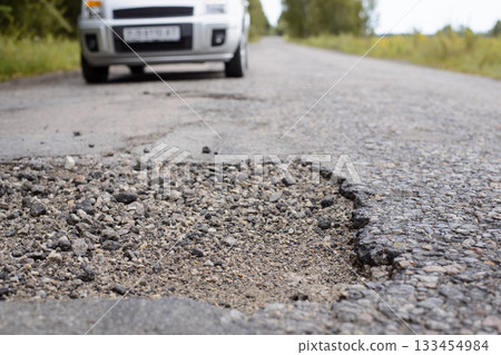 Country road in terrible condition. Modern grey vehicle approaching huge pothole Country road in terrible condition. Modern grey vehicle approaching huge pothole 133454984