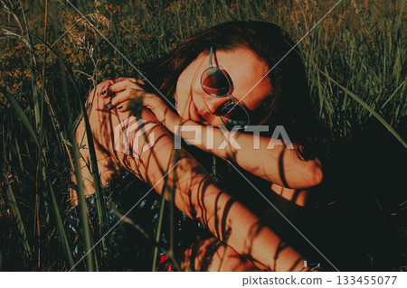 A woman wearing sunglasses relaxing in a summer field amongst tall grass on a sunny day, casting shadows on her skin. Moody female portrait in summer. 133455077