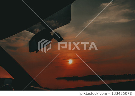 Sunset view through a car windshield in the evening. A beautiful sunset seen from inside an automobile. The orange and red hues of the sky create a stunning and peaceful atmosphere with a lake view. Sunset view through a car windshield in the evening. A beautiful sunset seen from inside an automobile. The orange and red hues of the sky create a stunning and peaceful atmosphere with a lake view. 133455078
