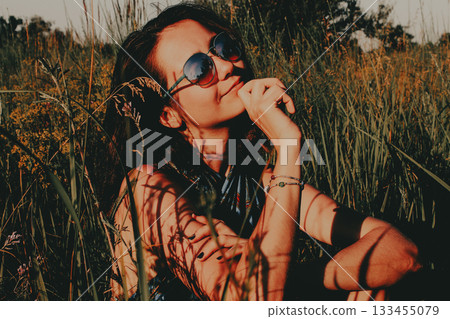 Young woman enjoying the sunlight in a grassy field. A serene portrait of a pretty girl wearing sunglasses, sitting on a lawn. Moody portrait. 133455079