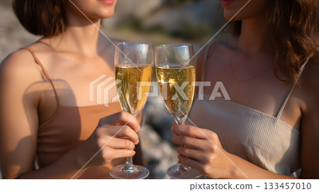 Two women celebrating with champagne glasses in a sunny outdoor setting by rocky landscape 133457010