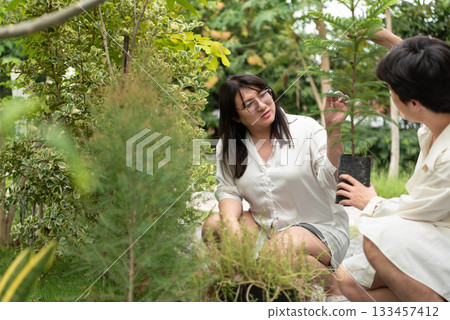 Two LGBTQ friends, one with long hair and the other with short hair, plant a tree in the morning garden. They share a joyful moment while caring for nature and nurturing new life in their home garden. Two LGBTQ friends, one with long hair and the other with short hair, plant a tree in the morning garden. They share a joyful moment while caring for nature and nurturing new life in their home garden. 133457412