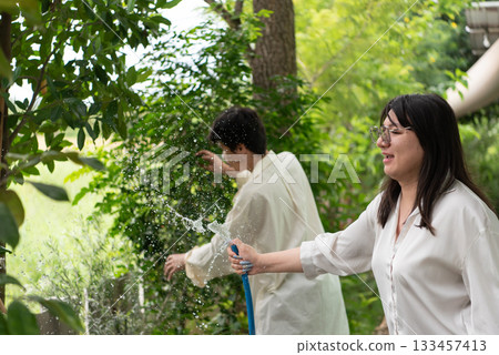 Two LGBTQ friends, one with long hair and the other with short hair, enjoy watering their garden in the morning. They share a joyful moment while caring for their plants in the lush outdoor space. 133457413