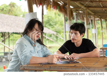 Two LGBTQ friends sitting at an Isaan Thai restaurant, enjoying their lunch together. One is writing notes while the other listens attentively, sharing a relaxed and enjoyable meal in cozy atmosphere. 133457445