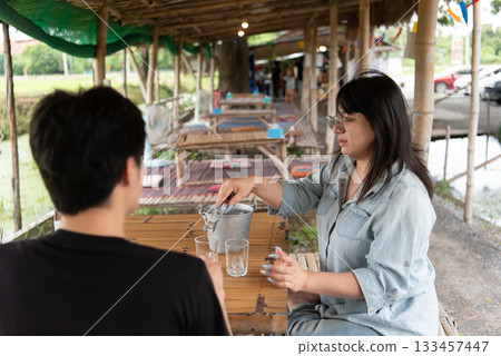 Two LGBTQ friends enjoying their lunch at an Isaan Thai restaurant. One is serving ice cubes into glasses while the other is seated, sharing a moment of connection in a cozy, vibrant atmosphere. 133457447