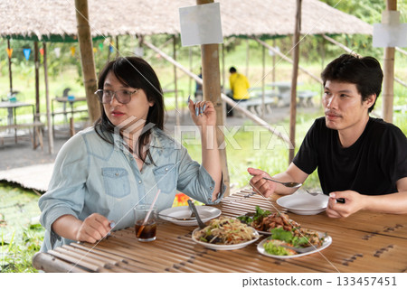 Two LGBTQ friends enjoying lunch at an Isaan Thai restaurant. One is holding a plate of food while the other watches, both sharing a moment of connection, laughter, and enjoyment in a casual setting. 133457451