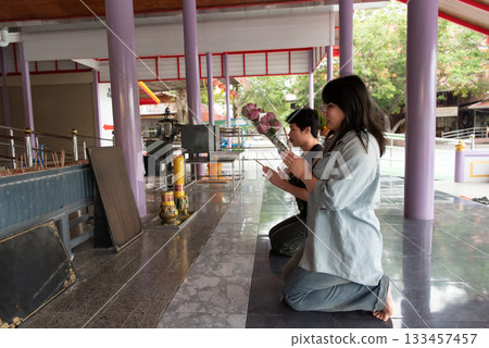 Two LGBTQ friends kneeling and praying at a Thai temple. One is holding flowers, while the other is in a peaceful prayer posture. They are performing a spiritual ritual in a tranquil temple setting. 133457457