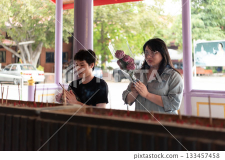 Two LGBTQ friends offering incense and flowers in prayer at a Thai temple. They are performing a spiritual ritual with devotion and respect in a peaceful temple environment. Two LGBTQ friends offering incense and flowers in prayer at a Thai temple. They are performing a spiritual ritual with devotion and respect in a peaceful temple environment. 133457458