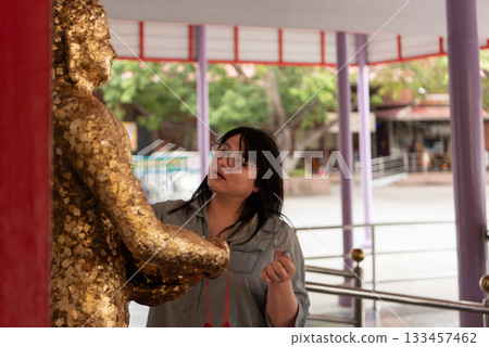 Two LGBTQ friends are performing a religious ritual at a Thai temple, offering flowers and paying respect by applying gold leaf to a Buddha statue as part of their spiritual practice. 133457462