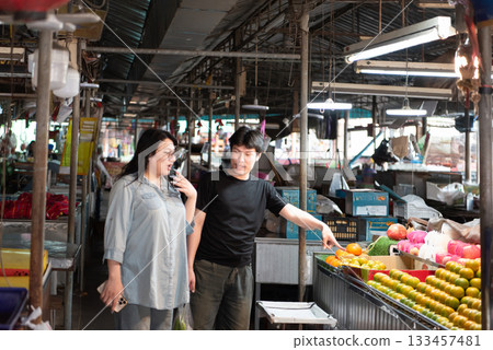 Two LGBTQ friends shopping for fresh vegetables at a local Thai market. One is pointing at the colorful fruits and vegetables, while the other is selecting produce, showing their lively interaction 133457481