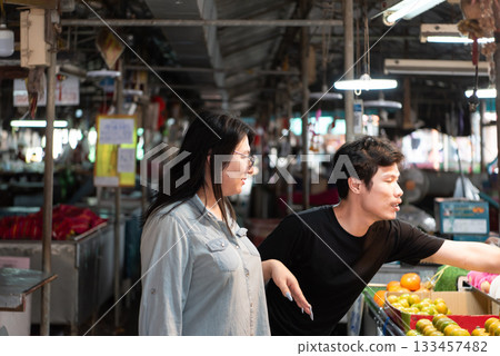 Two LGBTQ friends shopping for fresh fruits at a busy Thai market. One is pointing towards the produce, while the other is holding a cucumber, both actively engaged in the market vibrant atmosphere. 133457482