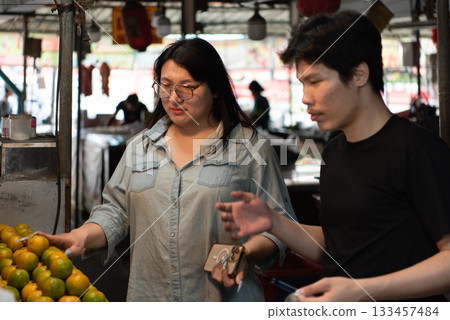 Two LGBTQ friends shopping for fresh fruits at a Thai market. One of them selects oranges while the other is holding a basket. The vibrant atmosphere of the busy market fills the scene. 133457484