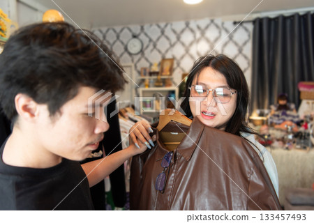 Two LGBTQ friends enjoy shopping for fashionable clothes at a boutique. One friend holds up a stylish leather jacket while the other discusses it, surrounded by trendy fashion items in a boutique. 133457493